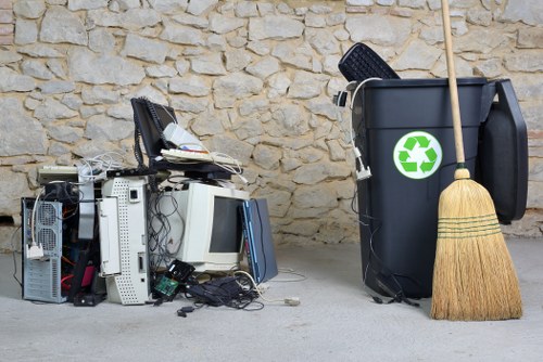 A collection of discarded electronic waste including old desktop computers, monitors, keyboards, and tangled cables, situated on a concrete surface in front of a rough-textured stone wall. To the right, a black wheelie bin with a green recycling symbol is positioned next to a traditional straw broom with a wooden handle, leaning against the bin. The electronic waste appears disorganized, with some components partially stacked and others scattered, indicative of a recent rubbish removal or garden clearance process. The environment appears to be an outdoor area possibly adjacent to a residential or commercial property in Feltham, suitable for rubbish collection services provided by Garden Clearance Feltham. The scene is well-lit with natural light, emphasizing the contrast between the electronic waste and the stone backdrop, highlighting the importance of proper rubbish disposal and recycling efforts in the local area.