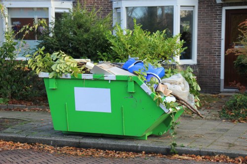 A large, green industrial skip situated on a paved driveway outside a residential property, filled with various discarded garden and household waste such as leafy branches, white cardboard boxes, blue plastic bags, and other miscellaneous rubbish. The skip is positioned close to the front of the house, with a brick wall, windows, and a partially visible door in the background. Surrounding the area are bushes, small trees, and a pathway with fallen leaves, indicating a garden clearance scene. The skip appears to be part of a garden clearance service in Feltham, with waste materials stacked towards the top edge, ready for removal by Garden Clearance Feltham. The lighting suggests a clear day, and the scene is typical of rubbish disposal activities in a residential neighbourhood with a focus on garden waste collection and clearance.