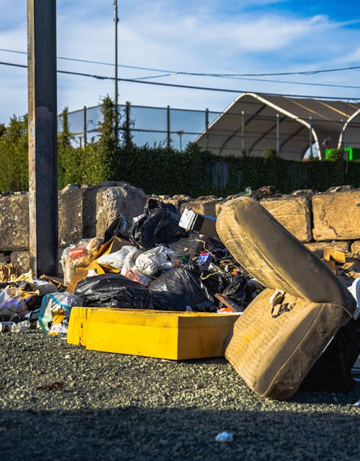 A large collection of mixed waste and rubbish bags, including cardboard boxes, plastic bottles, paper debris, and discarded packaging, is piled up on a paved sidewalk near a parking area. The waste in