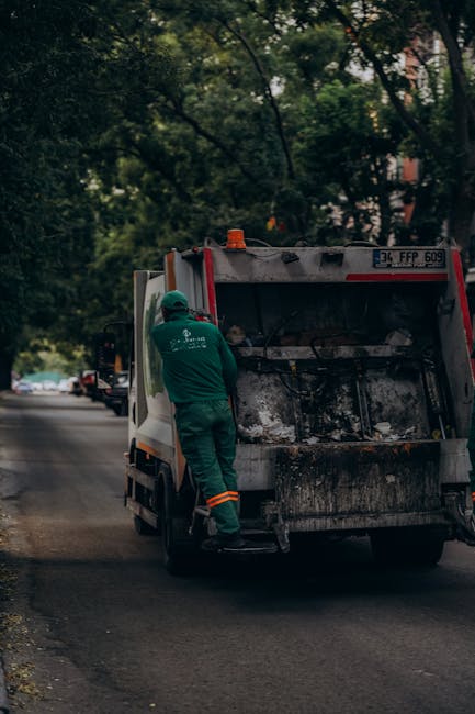 A waste collection worker dressed in green work uniform and protective helmet is emptying a black and white rubbish bin mounted on the back of a small commercial vehicle, positioned on a quiet urban s
