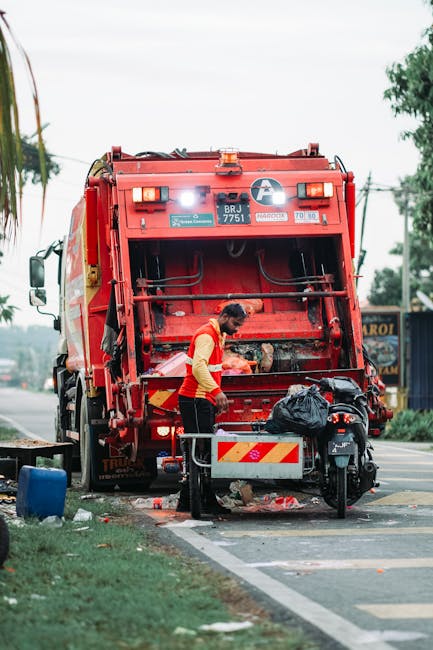 A waste collection worker dressed in green work uniform and protective helmet is emptying a black and white rubbish bin mounted on the back of a small commercial vehicle, positioned on a quiet urban s